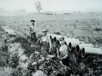 Three people stand in a muddy field with piping behind them. It is clearly an old photo in black and white with clothing from the early 1900s